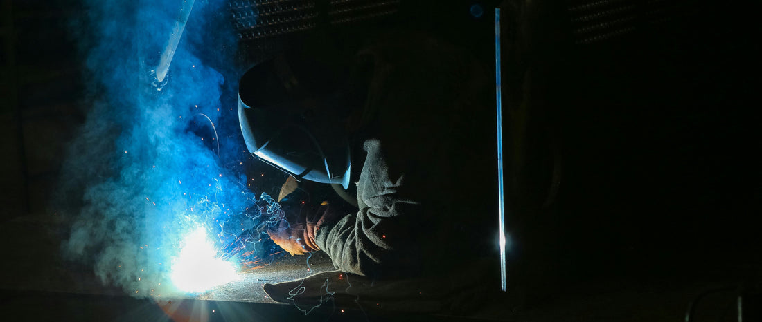 Welder wearing protective gear with bright sparks and blue welding smoke rising in a dimly lit workspace
